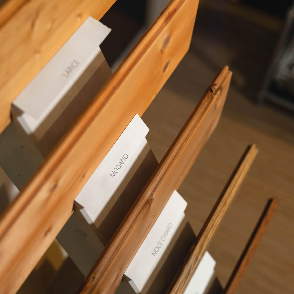 Dettaglio dall'alto di campioni di legno contrassegnati dalle etichette Larice, Mogano e Noce Chiaro in un espositore showroom Costrusion Len a Pordenone